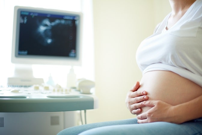 mulher grávida com ferropenia, com as mãos na barriga, monitor de USG ao fundo, em consulta de rotina
